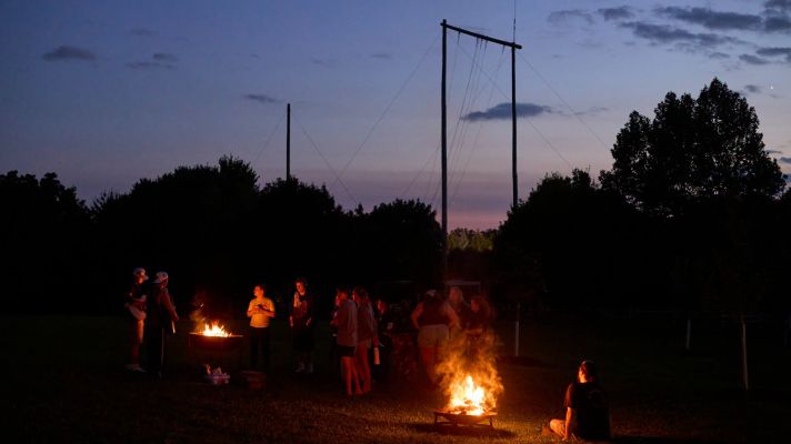 Young adults standing around two firepits in the early evening