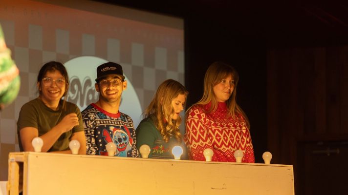 Four young adults standing at a game show style table wearing Christmas attire