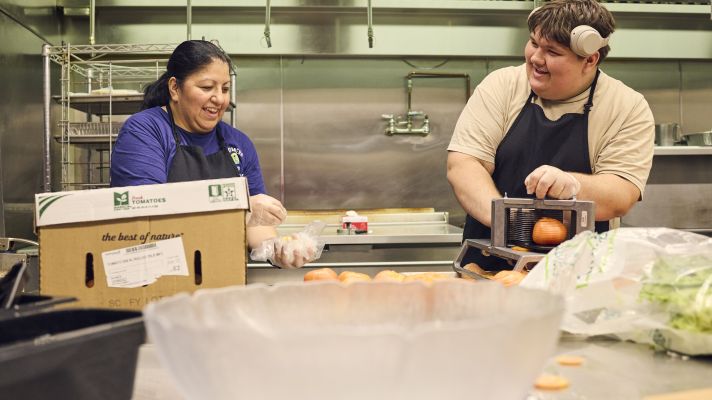 Adult female and young adult male laugh while preparing vegetables in an industrial kitchen