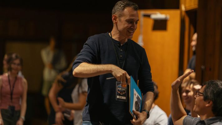 Middle aged male with a folder tucked under his forearm, looking at a teen sitting to his left